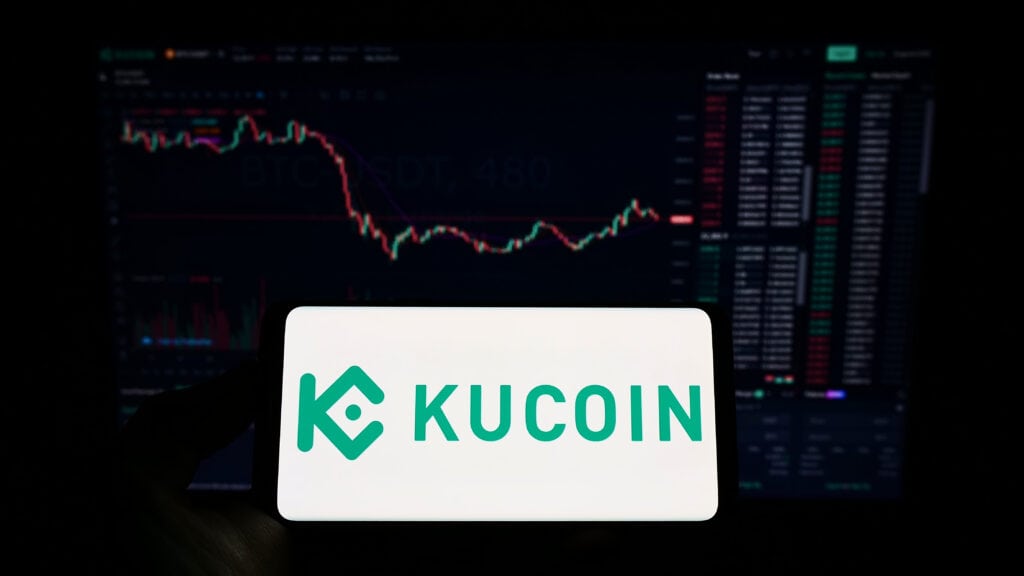 Person holding cellphone with logo of crypto exchange company KuCoin on screen in front of business webpage.