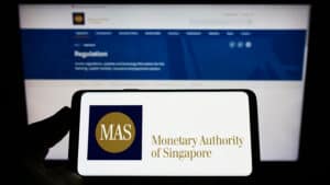 Person holding mobile phone with logo of Monetary Authority of Singapore (MAS) on screen in front of web page.