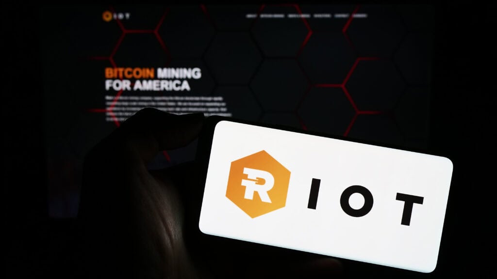 Person holding cellphone with logo of American Bitcoin mining company Riot Platforms Inc. on screen in front of webpage.
