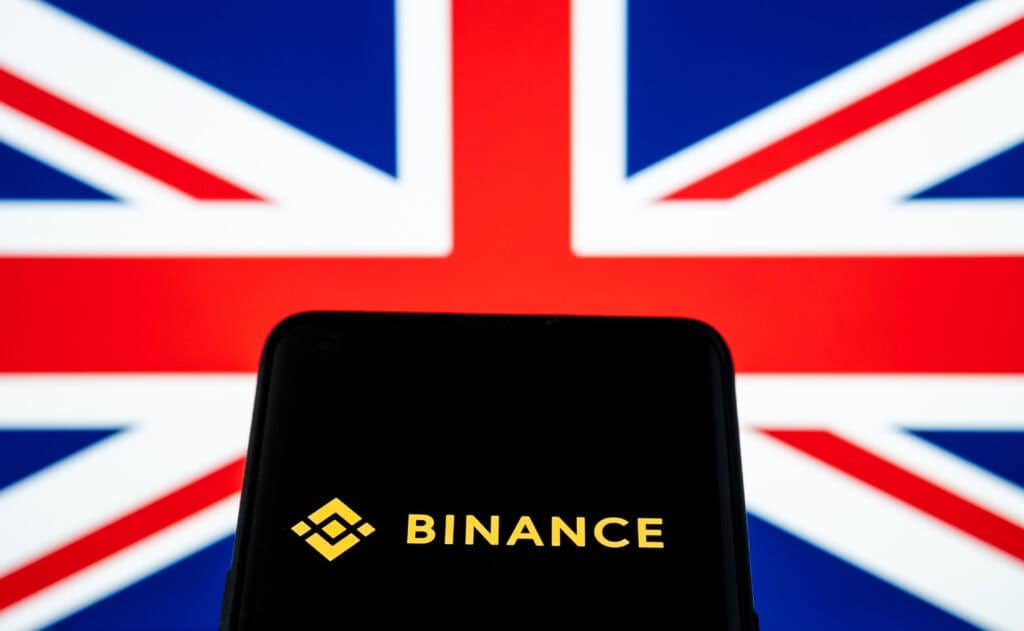 Binance logo seen on smartphone and blurred UK flag on the background.